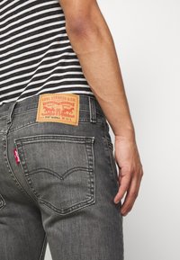 Grå denimjeans med en läderetikett på bakfickan, med sömnadsdetaljer och en liten röd tab. Randig skjorta synlig i bakgrunden.