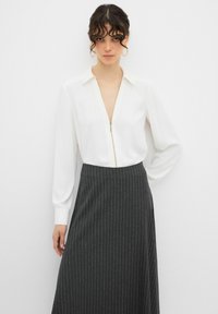 Blusa bianca con zip frontale e maniche lunghe, abbinata a una gonna scura a righe. Tessuto liscio e vestibilità su misura.