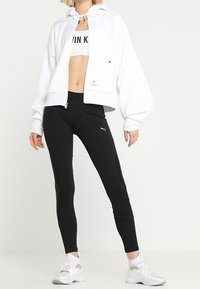Sweat à capuche blanc court avec fermeture éclair complète, poignets élastiques et capuche à cordon, associé à des leggings noirs taille haute et des chaussures de sport blanches.
