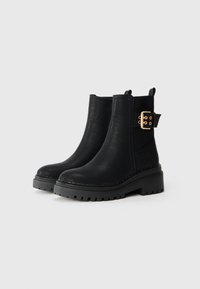 Bottines noires avec une finition en cuir texturé, des panneaux latéraux élastiques, une semelle épaisse et un accent en boucle dorée sur le côté.