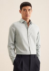 Camicia verde chiaro con maniche lunghe, colletto classico e colletto interno dettagliato. Abbinata a pantaloni scuri su misura.
