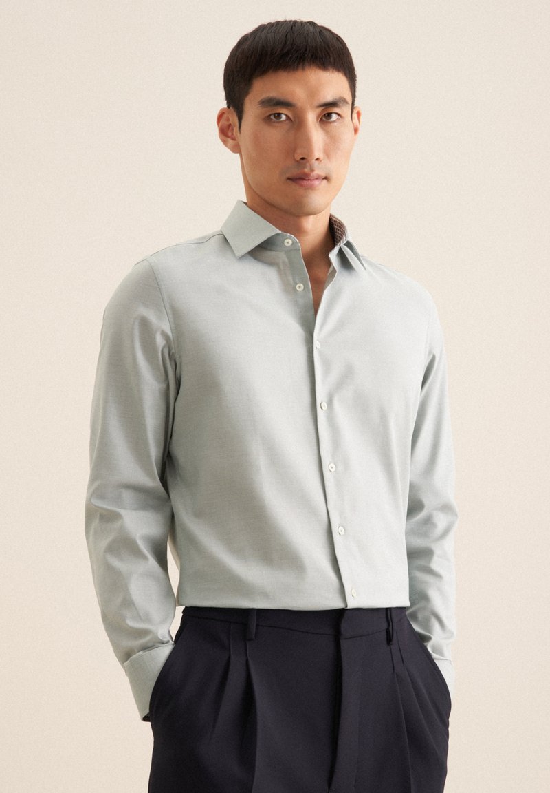 Camicia verde chiaro con maniche lunghe, colletto classico e colletto interno dettagliato. Abbinata a pantaloni scuri su misura.