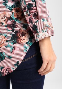 Blusa a fiori con righe rosse e crema, caratterizzata da un bordo arrotolato e un bottone marrone, con una texture di tessuto liscia.