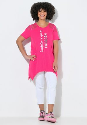Uśmiechnięta kobieta z kręconymi włosami, ubrana w jasnoróżową tunikę z napisem „FREEDOM”, białe spodnie oraz różowe sneakersy w panterkę.