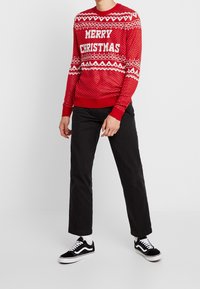 Rode gebreide trui met witte tekst "MERRY CHRISTMAS" en sneeuwvlokpatronen; gecombineerd met een zwarte broek en zwarte sportschoenen met witte accenten.