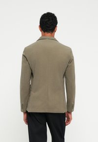 Olijfgroene blazer met een textuurstof, gestructureerde schouders, notch revers, enkele rugventilatie en vierknopen detail bij de mouwen.