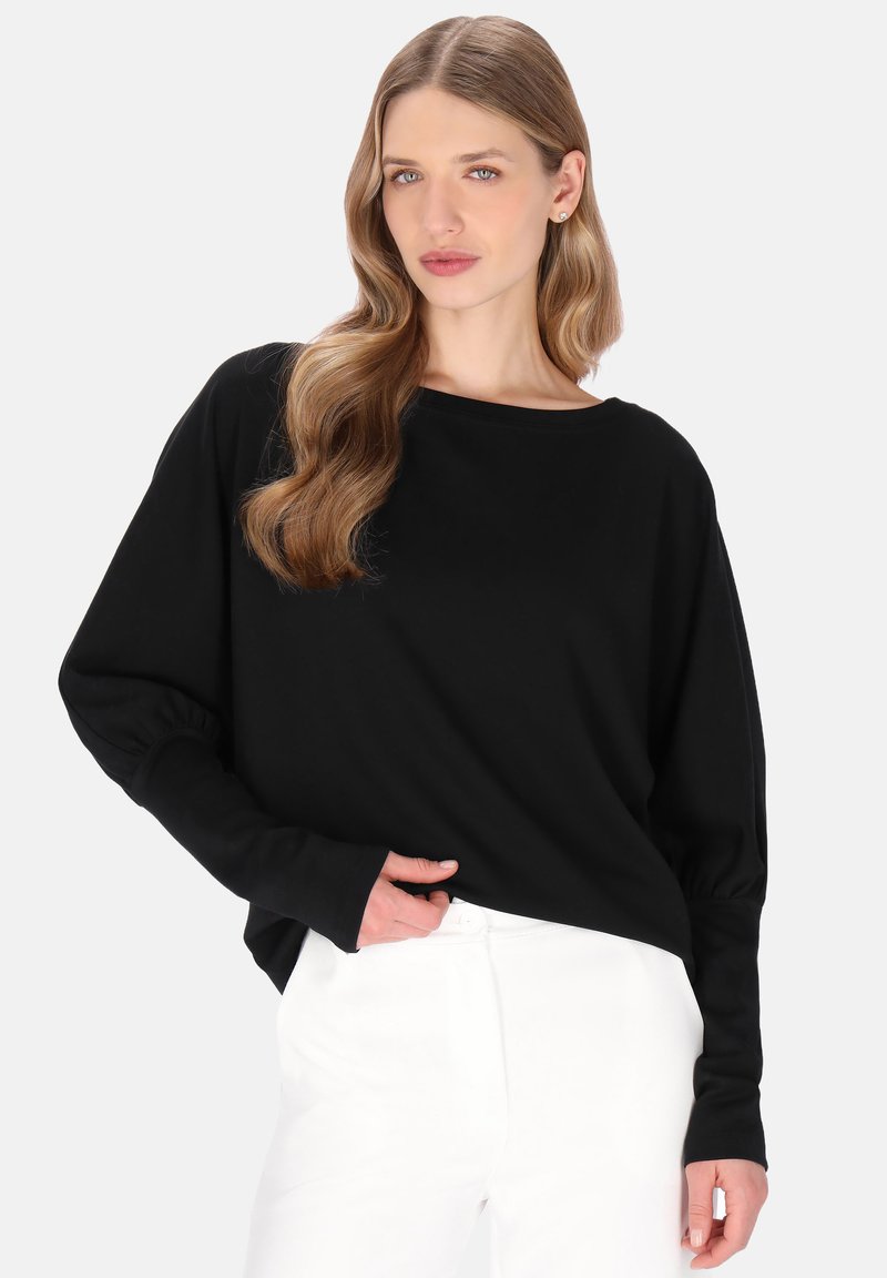 Haut noir à manches longues avec un large décolleté et une coupe ample. Tissu doux, manches froncées et ourlet arrondi contrastent avec un pantalon blanc.