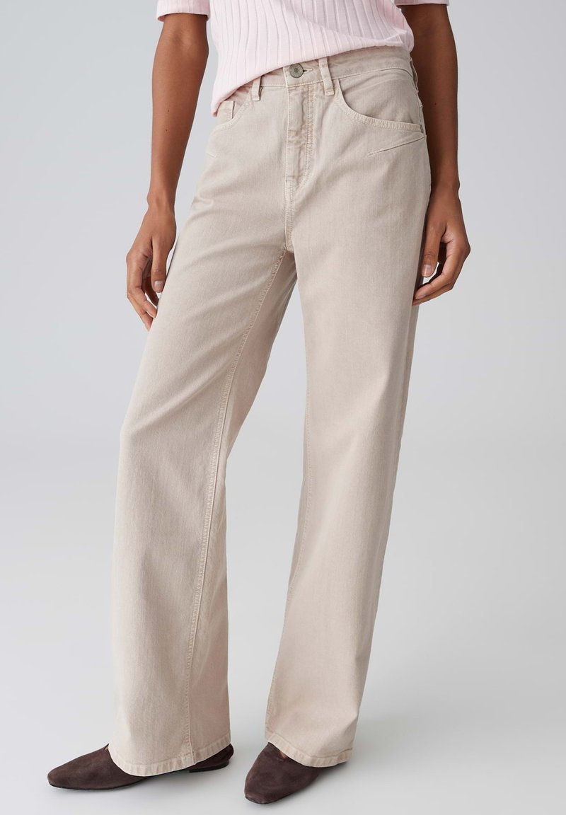 Donna che indossa jeans beige a gamba larga, top rosa a costine e scarpe marrone scuro con punta appuntita, in piedi davanti a uno sfondo grigio uniforme.