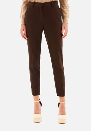 Person iført skræddersyede mørkebrune bukser til ankellængde og beige plateau højhælede sko, stående mod en ensfarvet baggrund.