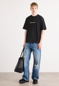 Svart bomull t-skjorte med hvit "Calvin Klein" logo, blå slitte jeans, svart lær totebag og kraftige svarte sko.