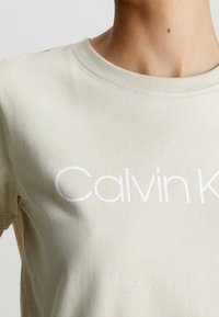 Ljus beige bomull t-shirt med rund halsringning, med en vit textlogotyp "Calvin K" placerad centralt på bröstet.