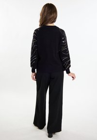Maglione nero a maniche lunghe con texture e dettagli metallici. Il corpo è completamente nero con polsini e bordo a coste. Abbinato a pantaloni neri a gamba larga.