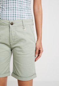 Světlé zelené šortky s ohrnutými lemy, se zapínáním na knoflík a háček a jemnou texturou. Kombinované s károvaným vzorem na topu.