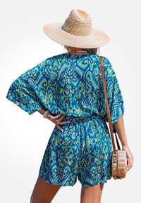 Frau in einem blau-grünen, gemusterten Jumpsuit, mit einem breiten Strohhut, die eine runde Korbtasche trägt und mit der Hand auf der Hüfte in die Ferne schaut.