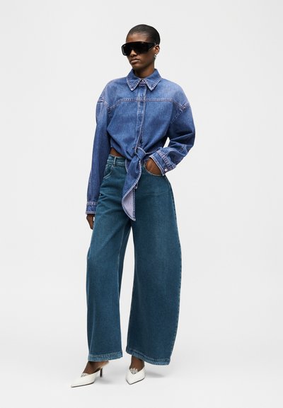 Μοντέλο που φοράει μπλε τζιν πουκάμισο μεγάλης διάστασης δεμένο στη μέση, φαρδύ μπλε τζιν παντελόνι, λευκά μυτερά τακούνια και μεγάλα μαύρα γυαλιά ηλίου σε απλό φόντο.