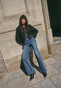 Svart texturerad bomberjacka, blå högmidjade utsvängda jeans och svarta spetsiga ankelboots, poserande mot en stenvägg som bakgrund.