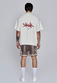 Chemise crème à manches courtes avec logo "SikSilk" rouge, assortie à un short brun brillant avec des bordures dorées, des chaussettes blanches et des baskets blanches.