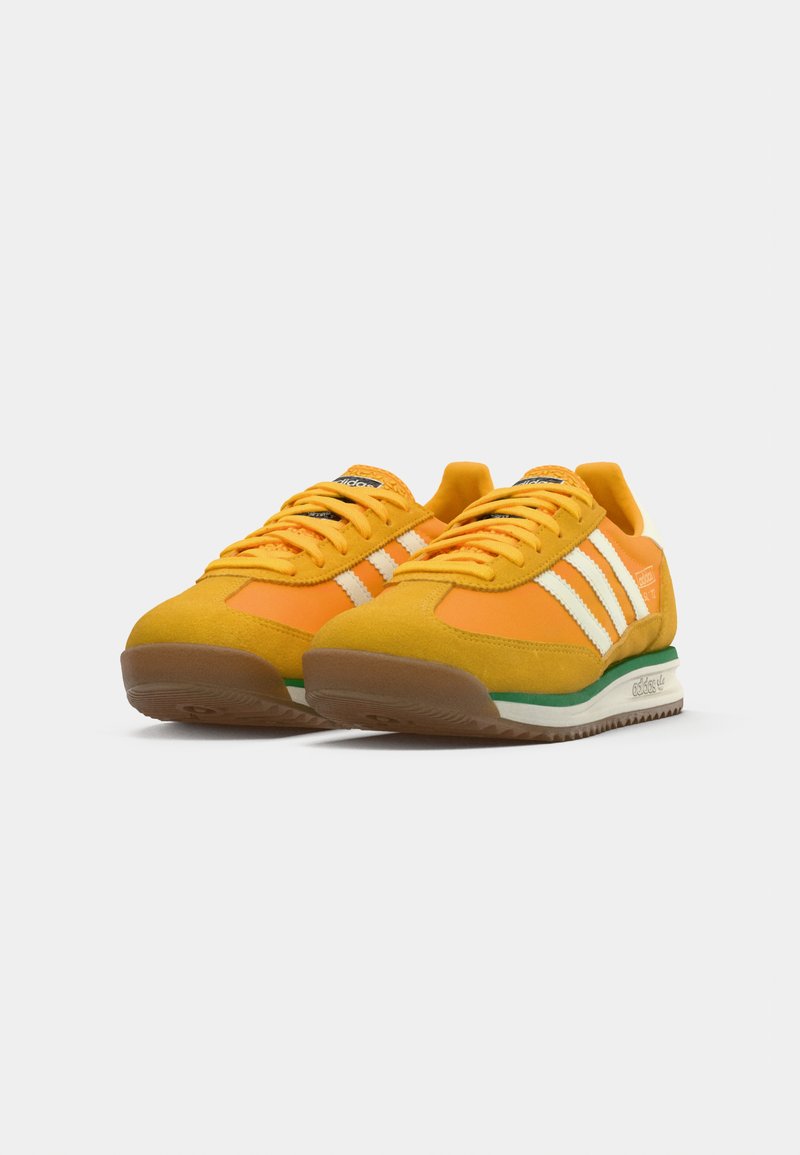 Un par de zapatillas Adidas amarillas y naranjas con rayas blancas, acentos verdes y suelas de goma sobre un fondo blanco.