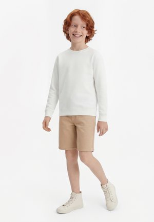 Enfant aux cheveux roux souriant, portant une chemise blanche à manches longues, un short beige et des baskets montantes blanches, debout avec un pied légèrement levé.