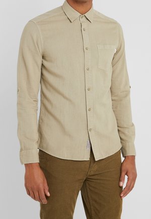 Man draagt een beige overhemd met lange mouwen en borstzak en een bruine broek, staand tegen een effen lichte achtergrond.