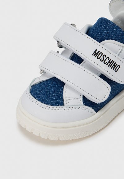 Μπλε τζιν και λευκό δερμάτινο πάνινο παπούτσι με δύο ιμάντες με βέλκρο και υφασμένο λαστιχένιο πέλμα. Το όνομα της μάρκας "MOSCHINO" είναι τυπωμένο με μαύρα γράμματα.