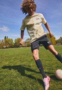 Heled kollane Nike t-särk logoga, mustad lühikesed püksid ja roosad jalgpallikontsadega. Mängija driblib jalgpalli rohelisel muruväljakul.
