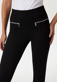 Black pants with a smooth texture, high waistband, and two zippered front pockets featuring silver hardware details.