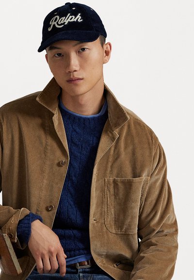 Ung mann i marineblå "Ralph" caps, brunt cordjacke og blå kabelstrikket genser, sitter mot en enkel hvit bakgrunn.