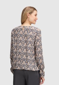 Blusa a maniche lunghe con un motivo geometrico nei toni del beige, grigio e blu. Il retro presenta una piccola apertura a forma di chiave. Tessuto morbido, vestibilità rilassata.