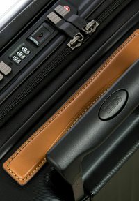 Valigia nera con superficie testurizzata, accentuazione e manico in pelle marrone a contrasto, doppie cerniere e lucchetto a combinazione.