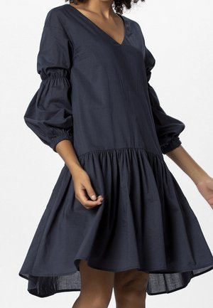 Woman wearing a loose navy blue V-neck dress with gathered long puff sleeves and a flared ruffled hem above the knees.