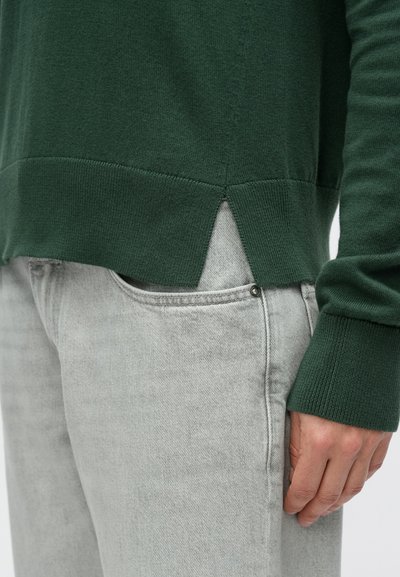 Σκούρο πράσινο πλεκτό πουλόβερ με ριμπ μανσέτες και σχισμή στο τελείωμα, συνδυασμένο με ανοιχτό γκρι τζιν. Λείος υφάδι, casual εφαρμογή.