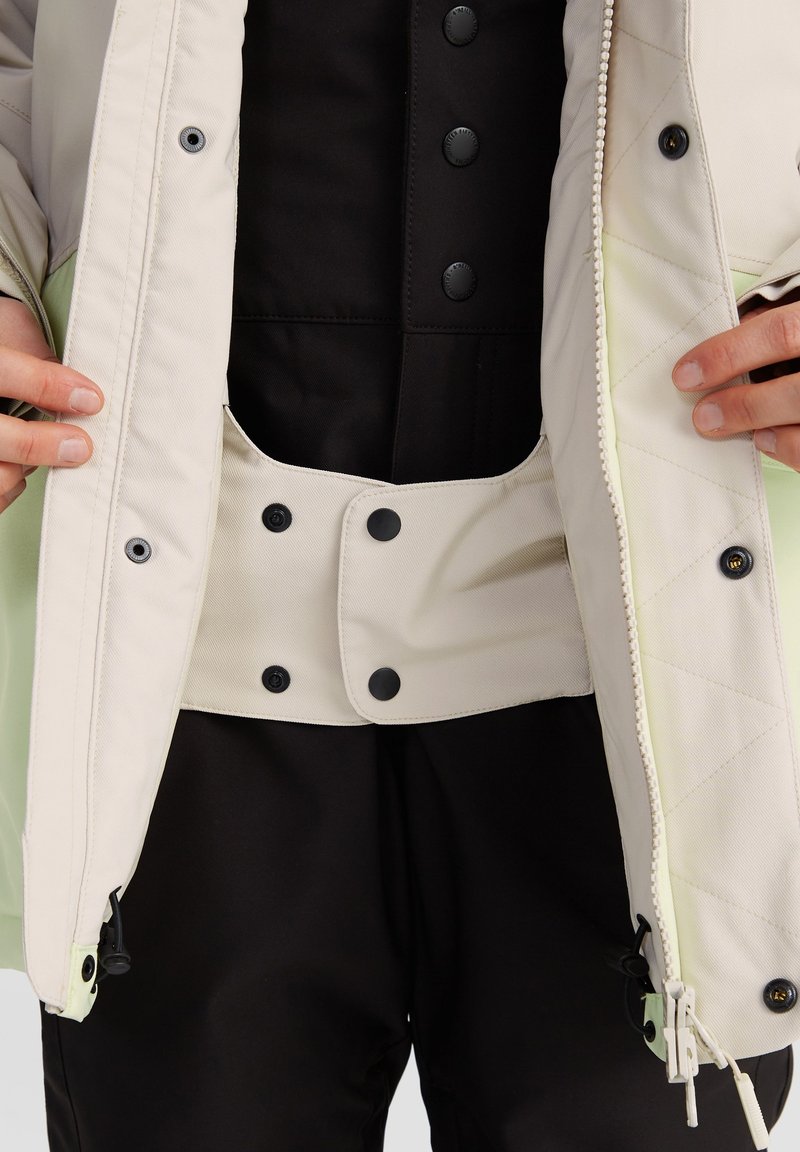 Jasno-kolorowa kurtka z czarnymi guzikami, pikowana tekstura; wewnętrzna warstwa czarna z kieszenią i zapięciem na nap; noszona na czarnych spodniach.