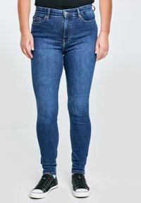 Osoba u tamnoplavim uskim trapericama i crnim tenisicama stoji naslonjena na jednobojnu svijetlu pozadinu.