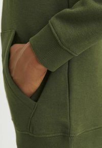 Felpa verde oliva con polsini a coste e tasca laterale. Il materiale sembra morbido con una texture liscia.