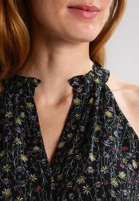 Dolna część twarzy i szyi kobiety w czarnej bluzce na ramiączkach z żółtymi, różowymi i białymi kwiatowymi wzorami oraz falbaniastego dekoltu.