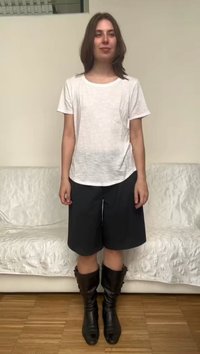 Jeune femme aux cheveux châtain mi-longs portant un t-shirt blanc, un short noir longueur genoux et des bottes noires montant jusqu'au milieu du mollet, debout à l'intérieur.