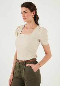 Haut beige côtelé avec décolleté carré, manches bouffantes et manches courtes. Porté avec un pantalon vert olive et une ceinture marron, mettant en valeur une silhouette ajustée.