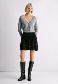 Cardigan grigio a maglia con chiusura a bottoni, gonna nera in tulle a strati e stivali neri in pelle al ginocchio. Design semplice e texture morbide.