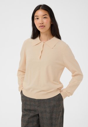 Femme aux longs cheveux noirs portant un pull texturé beige clair avec col et un pantalon à carreaux gris foncé, debout avec les mains dans les poches.