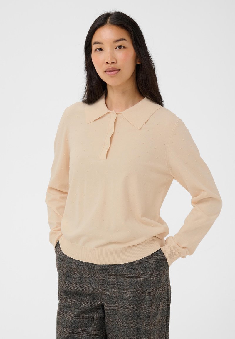 Femme aux longs cheveux noirs portant un pull texturé beige clair avec col et un pantalon à carreaux gris foncé, debout avec les mains dans les poches.