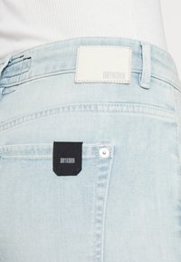 Vaqueros de mezclilla azul claro con una etiqueta de cuero blanco en la parte posterior de la cintura y una pequeña etiqueta negra en un bolsillo trasero, con costuras en contraste.