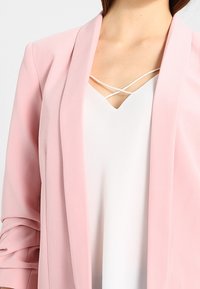 Světle růžový blazer s jemnou texturou, otevřeným předním dílem a ohrnutými rukávy, doplněný o bílý top s křížovým výstřihem.