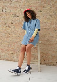 Jeansskjortklänning med korta ärmar, med knappfront. Bärs över en gulrandig långärmad skjorta, ihop med marinblå högskoor.