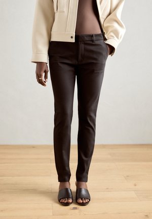 Personne portant un pantalon slim foncé, une veste beige et des sandales noires ouvertes aux orteils debout sur un sol en bois.
