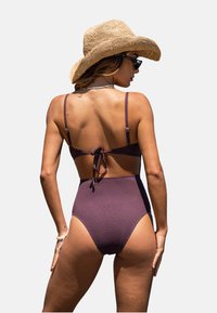 Bikini violet taille haute en tissu texturé, avec des bretelles fines réglables et un haut à nouer dans le dos. Accompagné d'un chapeau en paille.