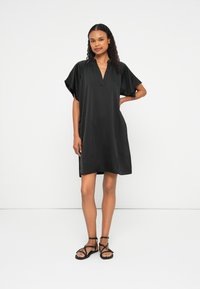 Vestido preto curto com decote em V e mangas largas e onduladas. Tecido suave e leve; comprimento até o joelho. Combinado com sandálias pretas de tiras.