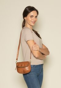 Sac bandoulière en cuir marron avec une forme arrondie, fermeture éclair et quincaillerie dorée. La bandoulière est ajustable et texturée.