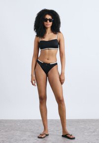Μαύριο μπικίνι δύο τεμαχίων με τοπ μπαντό που διαθέτει λεπτούς ιμάντες και σλιπ με χαμηλή μέση. Φορεμένο με μαύρα σαγιονάρες και γυαλιά ηλίου. Ομαλή υφή.