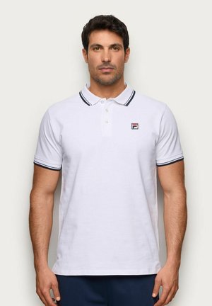 Hombre con polo blanco con ribete negro en el cuello y las mangas, de pie frente a un fondo claro y liso.